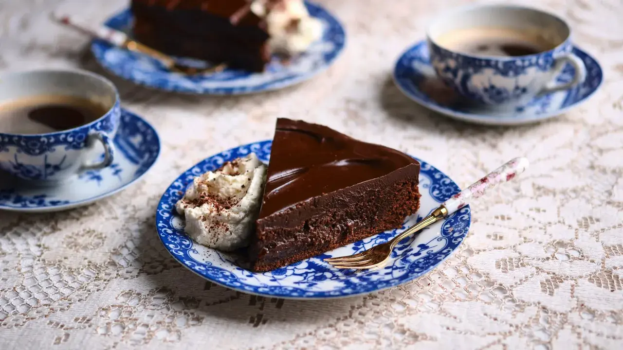 En bit kladdkaka med chokladganache och vispad grädde, serverad med kaffe på blåvita koppar.