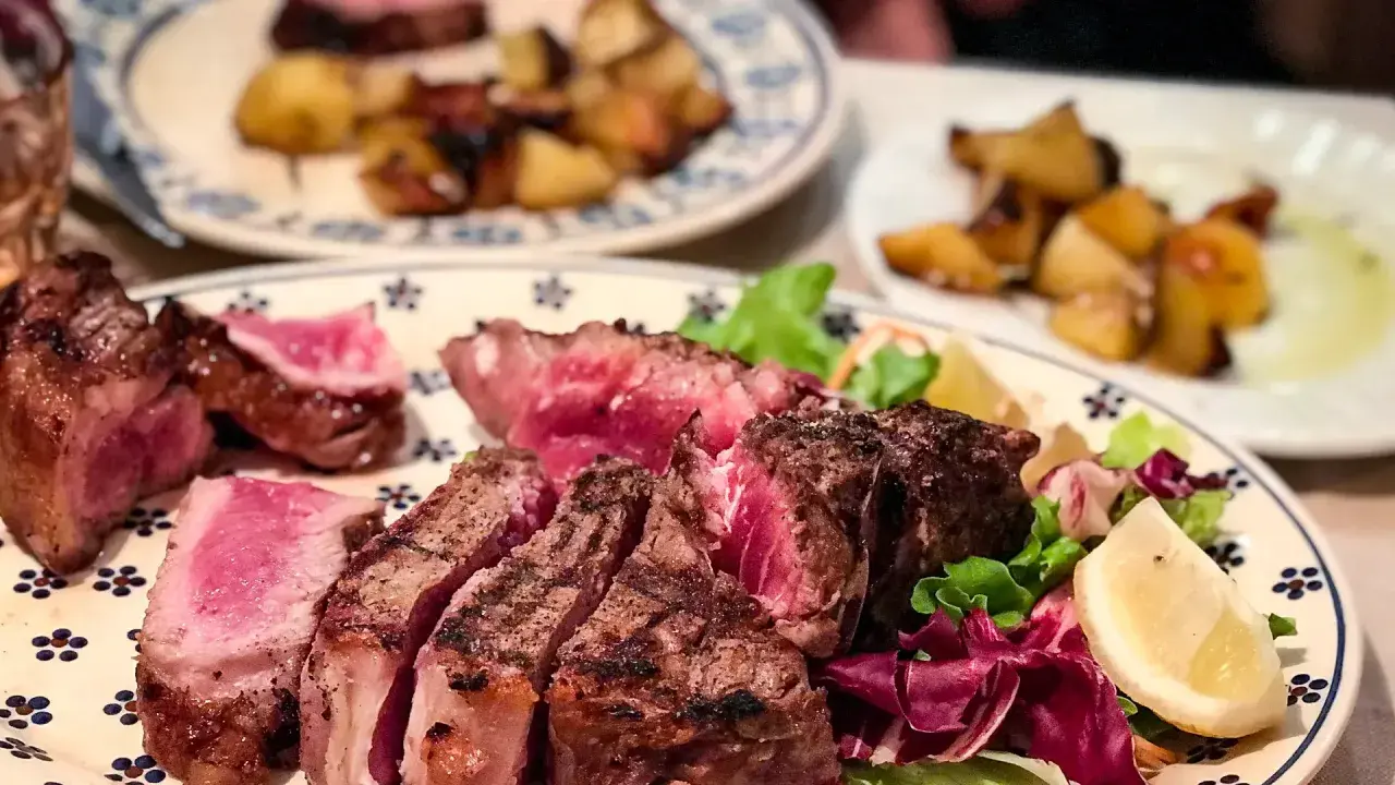 En saftig bistecca fiorentina, perfekt grillad med en rosa kärna, serveras med krispig sallad och citronklyfta. Rostade potatisar i bakgrunden.