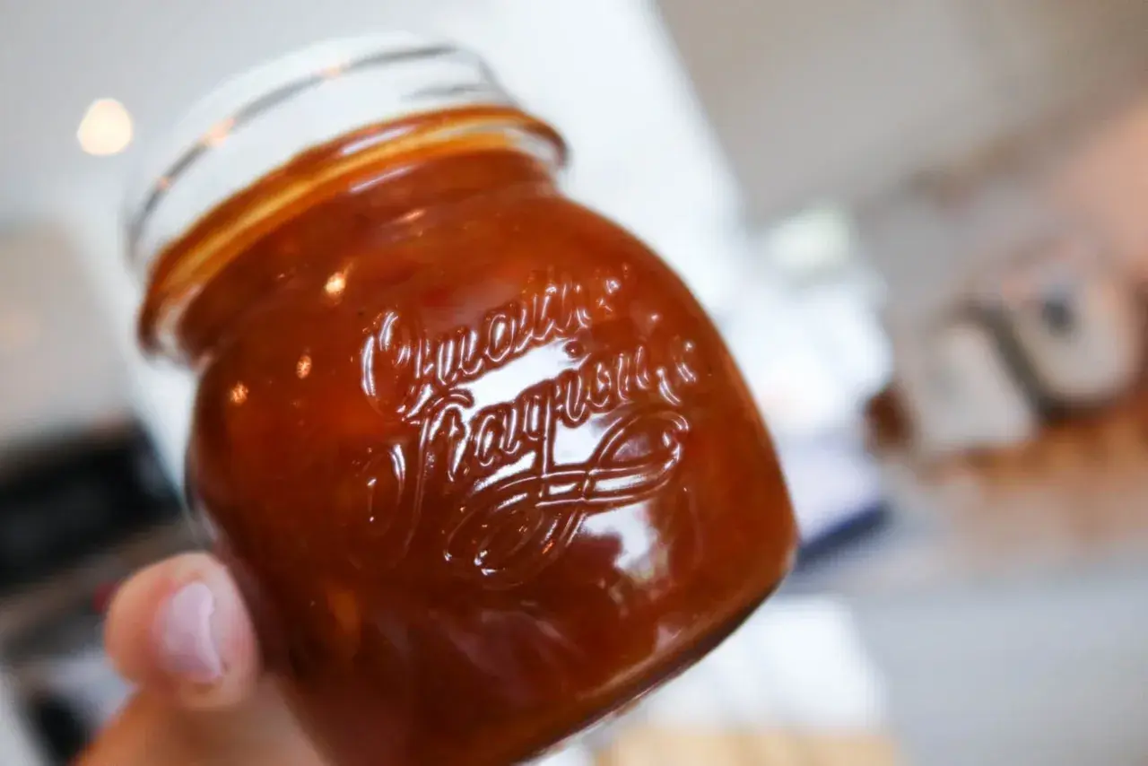 En hand håller en burk med hemlagad rabarberchutney. Den mörkröda chutneyn glänser i ljuset.