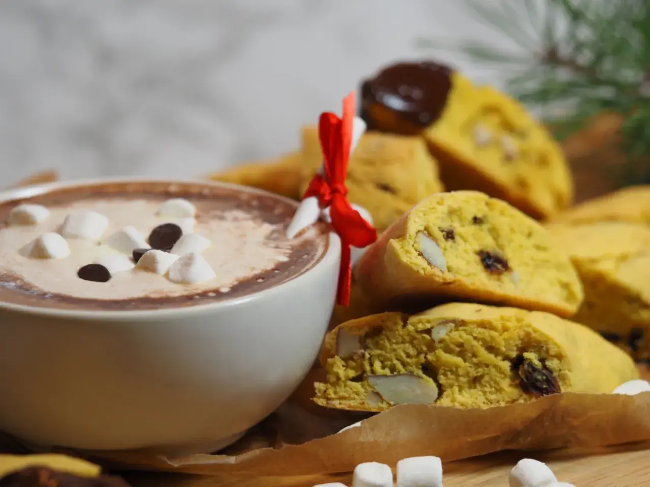 En kopp varm choklad med marshmallows och chokladbitar bredvid en hög med saffransbiscotti med mandel, inslagna i ett rött band.