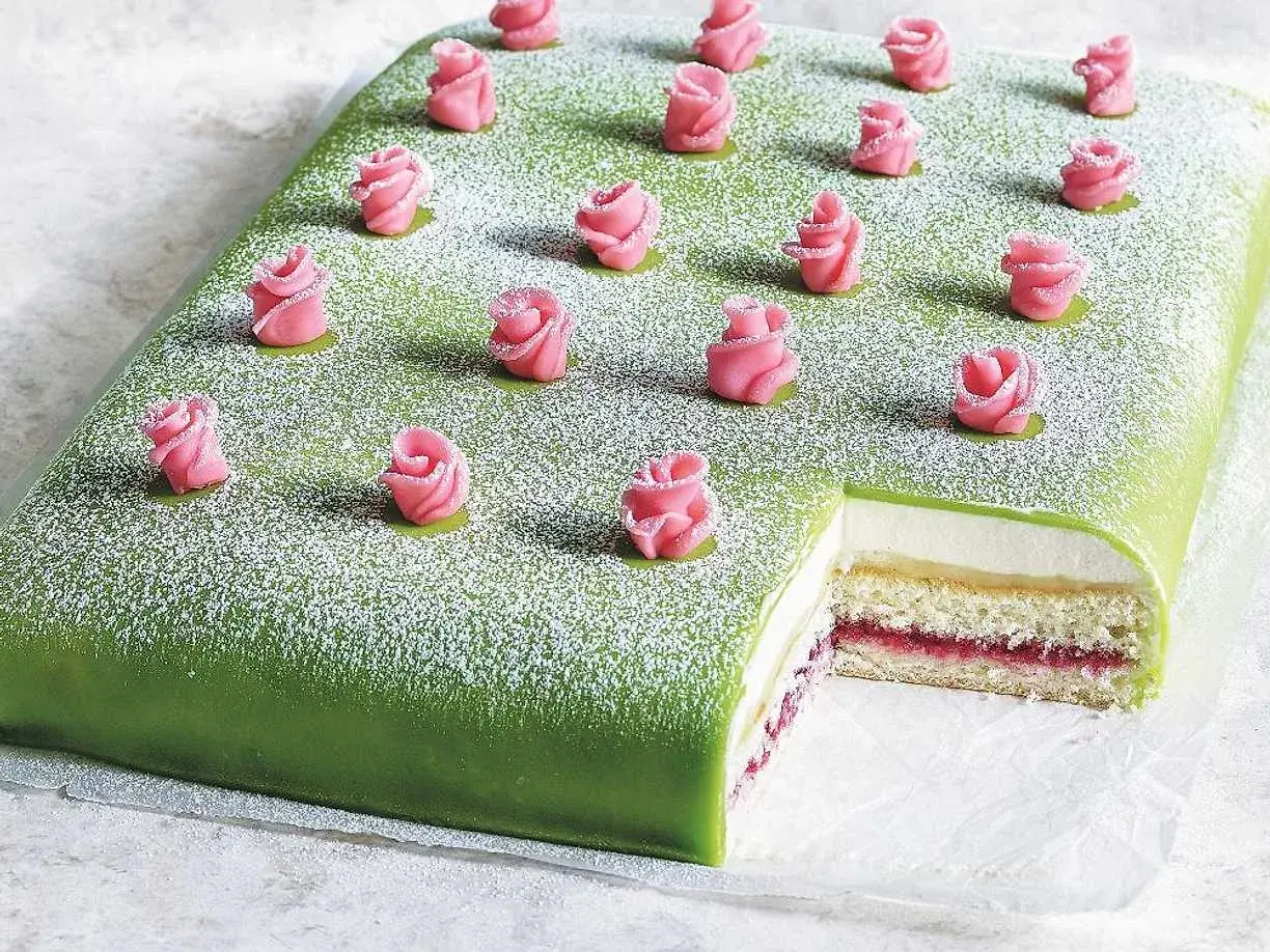 En grön täckt liten tårta med rosa marsipanrosor och pudersocker. En bit är bortskuren och visar lager av grädde och sylt.
