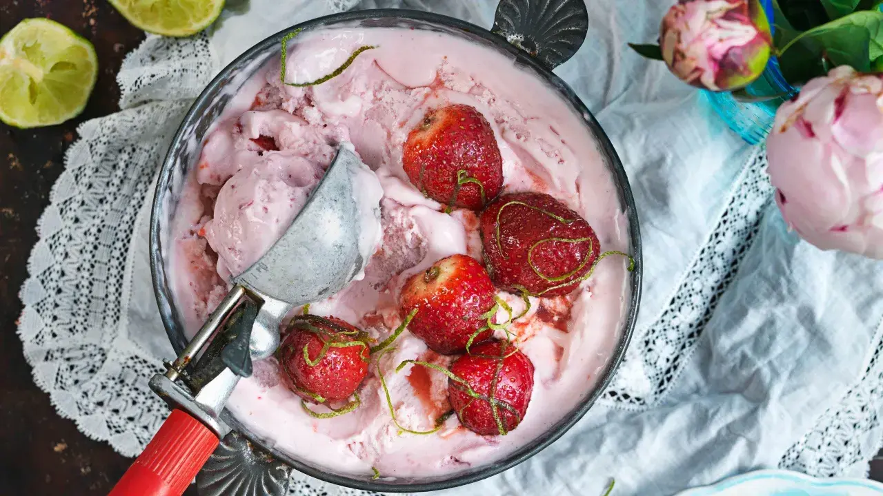 En skål med rosa jordgubbsglass, toppad med hela jordgubbar och limezest. En glasskopa vilar i skålen.