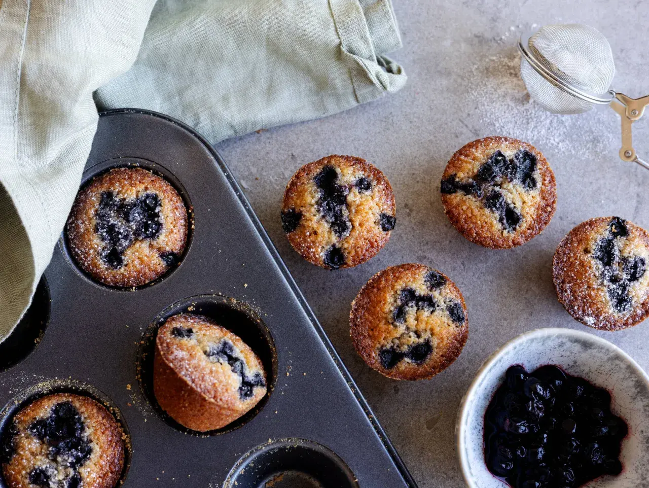 Nybakade blåbärsmuffins, en klassisk financier kaka, ligger i en form bredvid en skål med sylt och en sil.