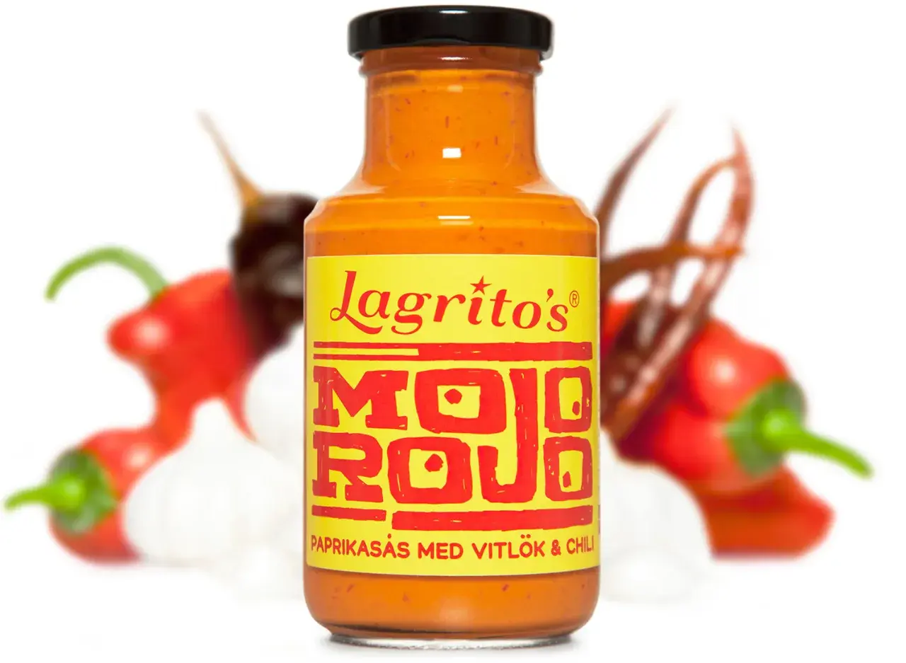 En flaska Lagrito's Mojo Rojo paprikasås med vitlök och chili, omgiven av suddiga röda chilifrukter och vitlöksklyftor.