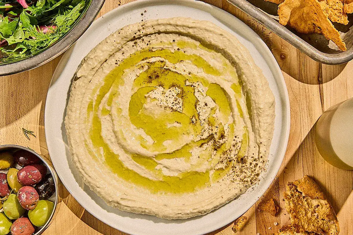 Krämig hummus med olivolja och kryddor, perfekt för ditt nästa recept. Serveras med oliver och pitabröd.