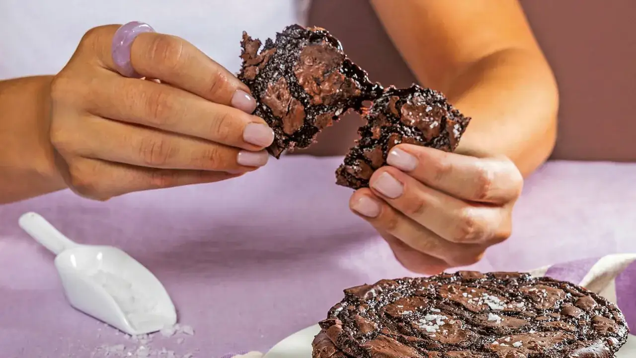 Händerna bryter isär en saftig chokladkaka med havssalt. En skopa med salt och fler chokladkakor syns i bakgrunden.
