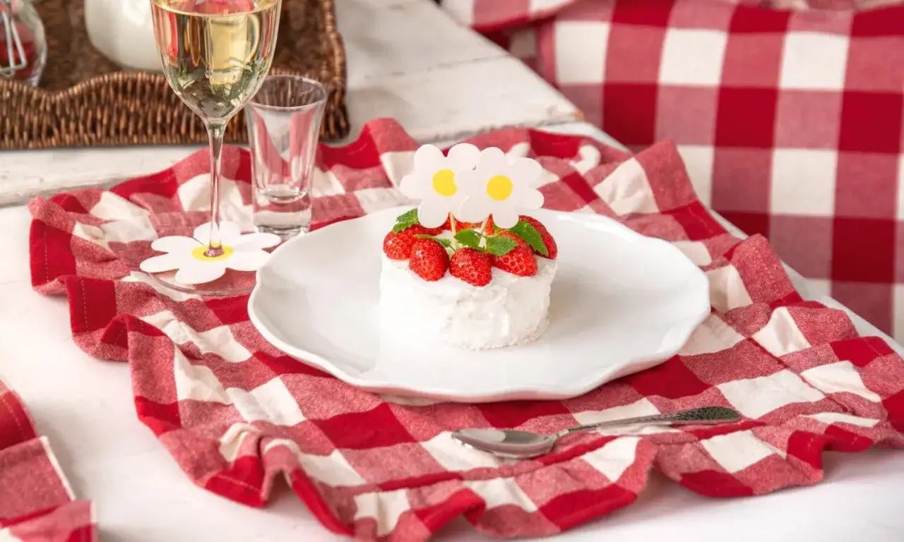 En liten, himmelsk jordgubbstårta med grädde och färska jordgubbar, dekorerad med blommor. Serveras på en vit tallrik med en rödvitrutig duk.