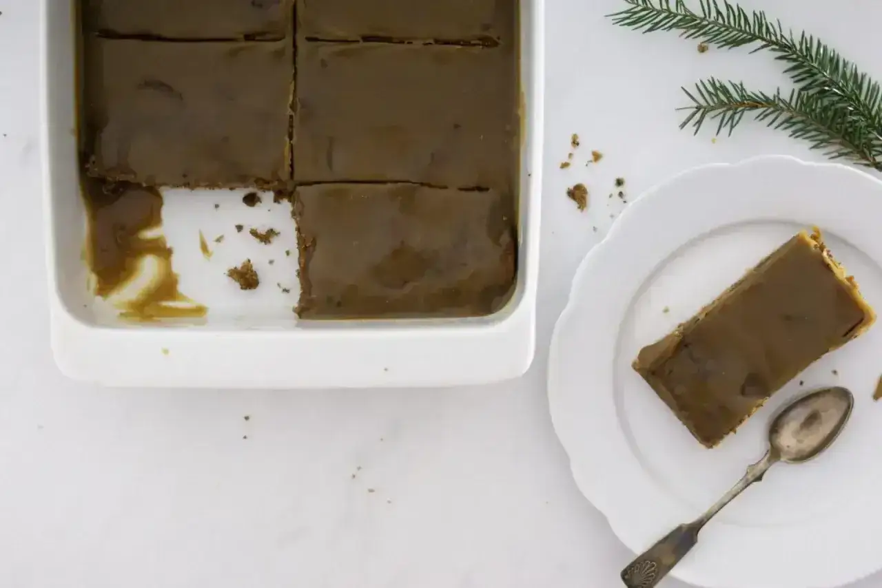En bit av en härlig sticky toffee pudding på en vit tallrik, med resten i en ugnsform. En grankvist ligger bredvid.