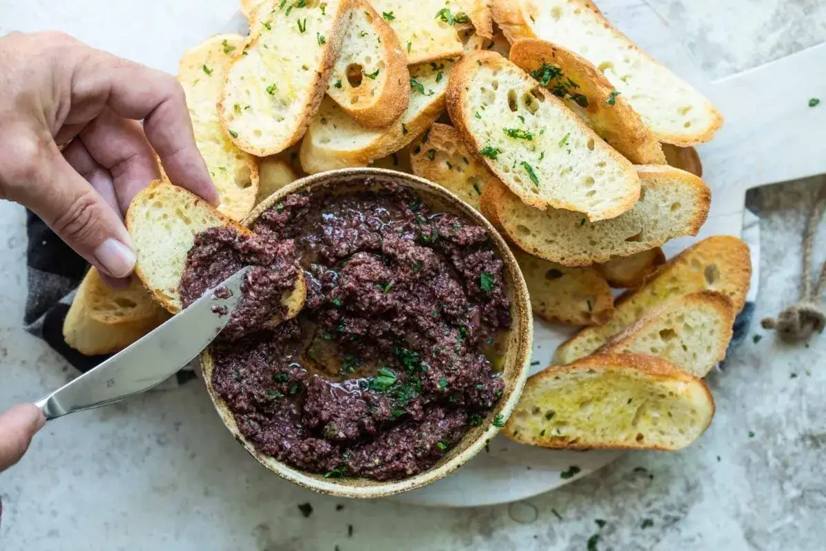 En hand breder ut en olivtapenade p&aring; en rostad br&ouml;dskiva. Ett enkelt och gott tapenade recept.