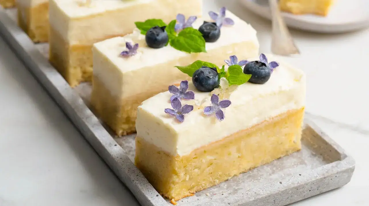 Små, eleganta bitar av ljus tårta med krämig topping, dekorerade med blåbär, mynta och vackra kanderade syrenblommor.