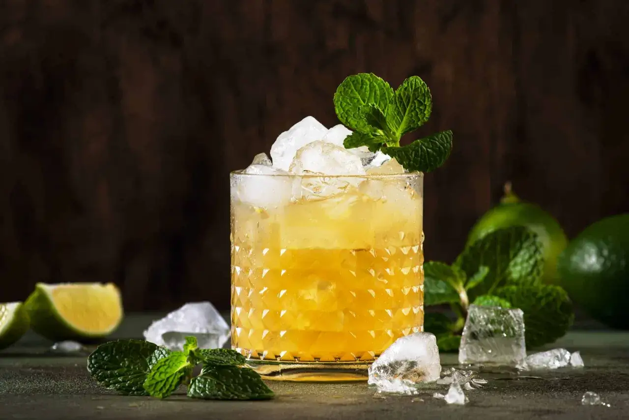 En gyllene Mai Tai-drink med is och mynta, omgiven av limeklyftor och isbitar.