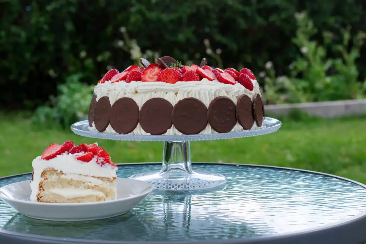 En vacker, stor jordgubbstårta med chokladdekorationer står på ett tårtfat. Bredvid ligger en bit av den lilla tårtan, redo att avnjutas i trädgården.