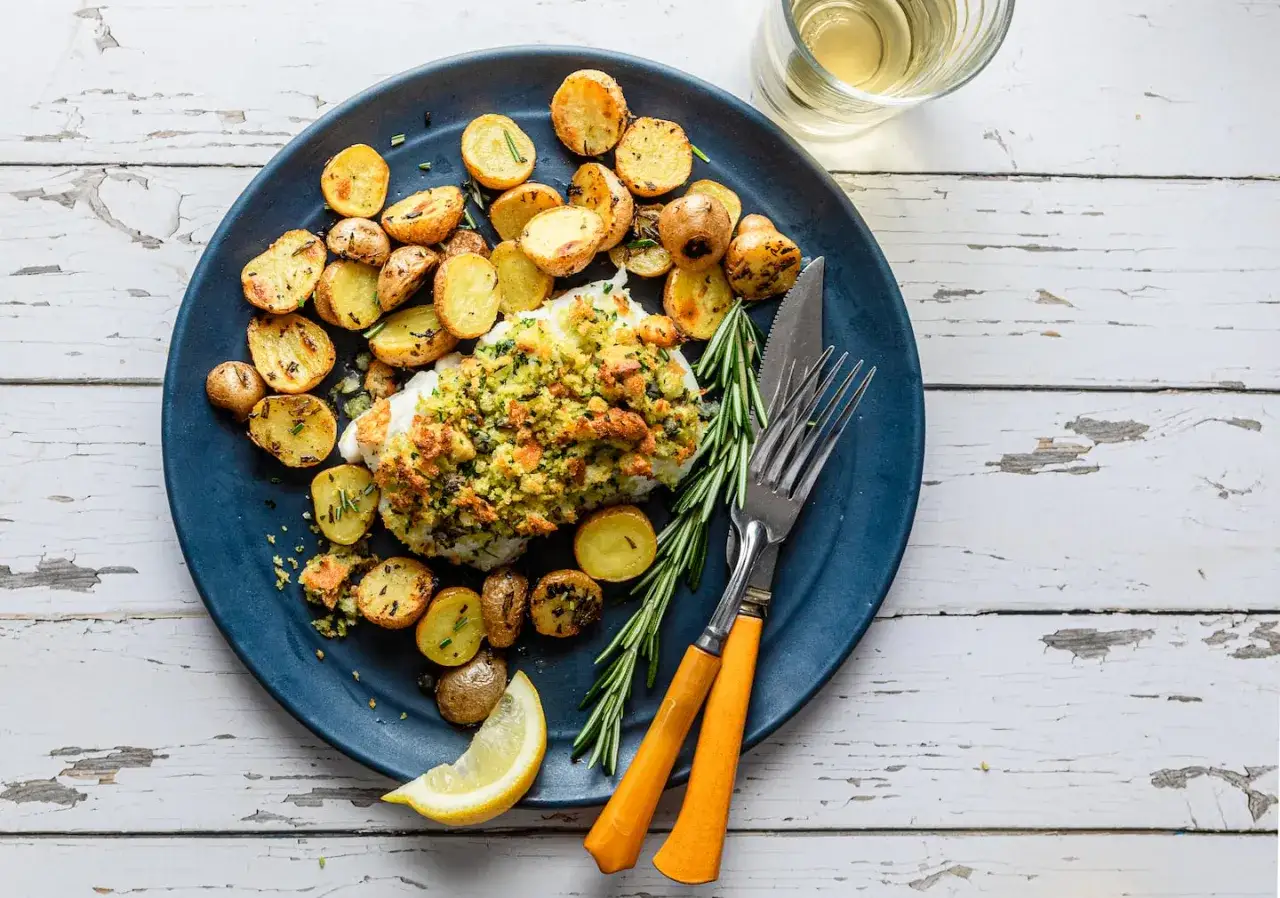 En härlig tallrik med torsk i ugn, toppad med krispigt ströbröd och örter, serverad med ugnsrostade potatisar och en citronklyfta.