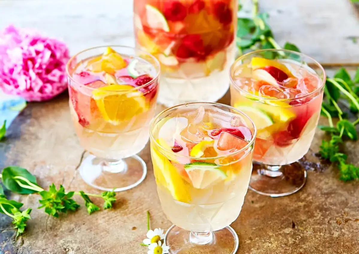 Tre glas med vit sangria cava fyllda med frukt och is. En stor karaff syns i bakgrunden.