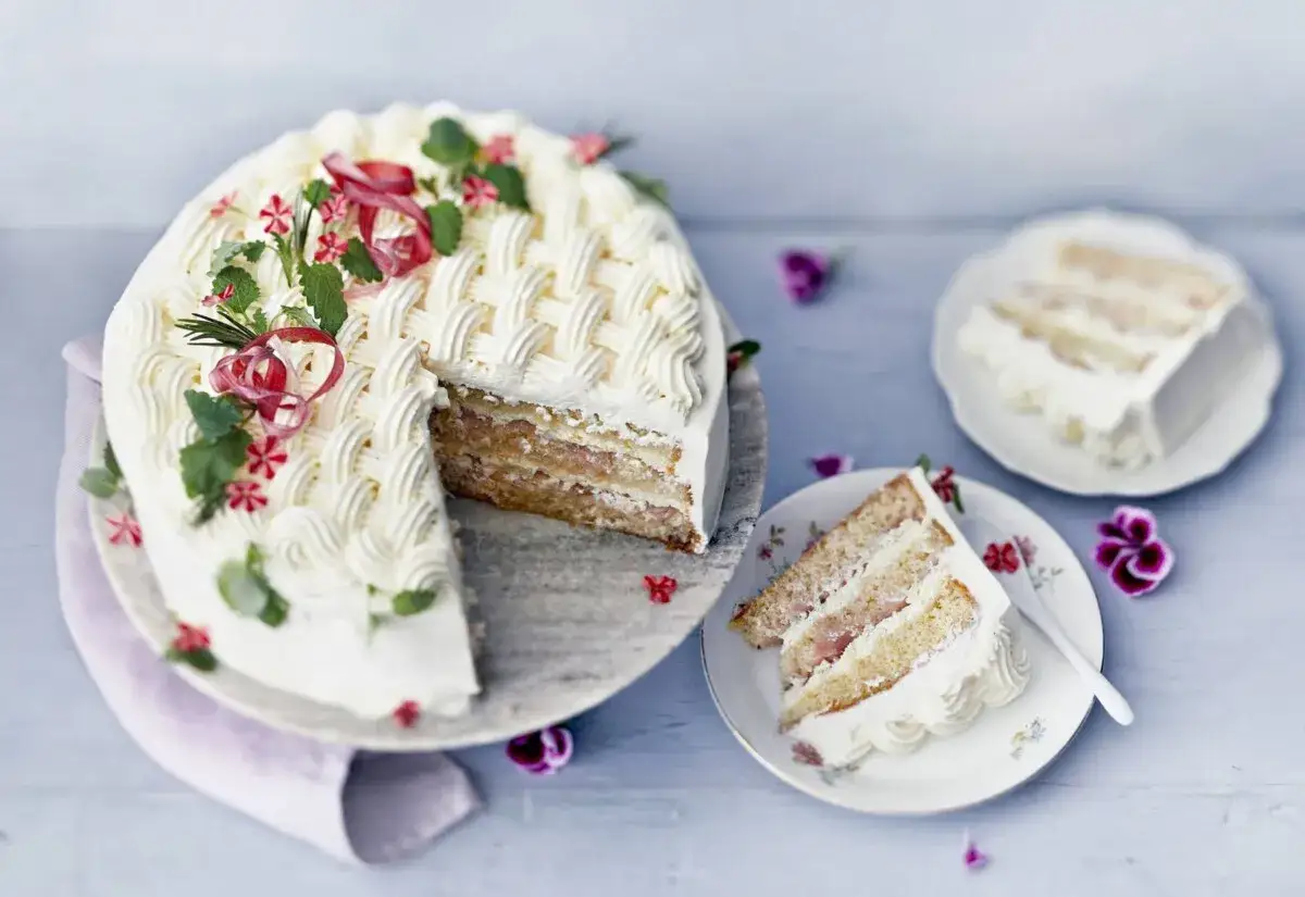 En vacker rabarberkompott tårta med flätmönster av grädde, dekorerad med örter och blommor. En bit är upplagd på en tallrik.