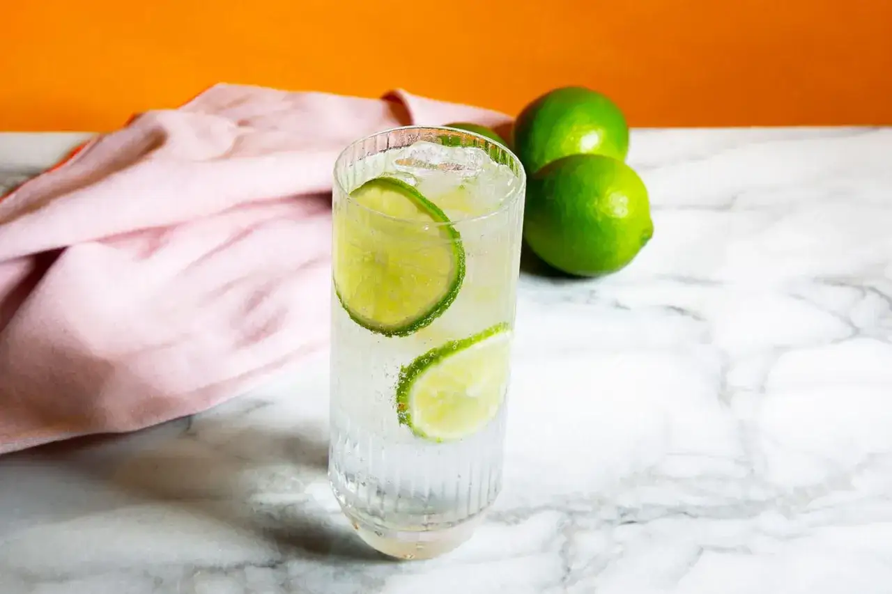 Ett glas gin och tonic med is och lime, bredvid tre hela limefrukter och en rosa duk.