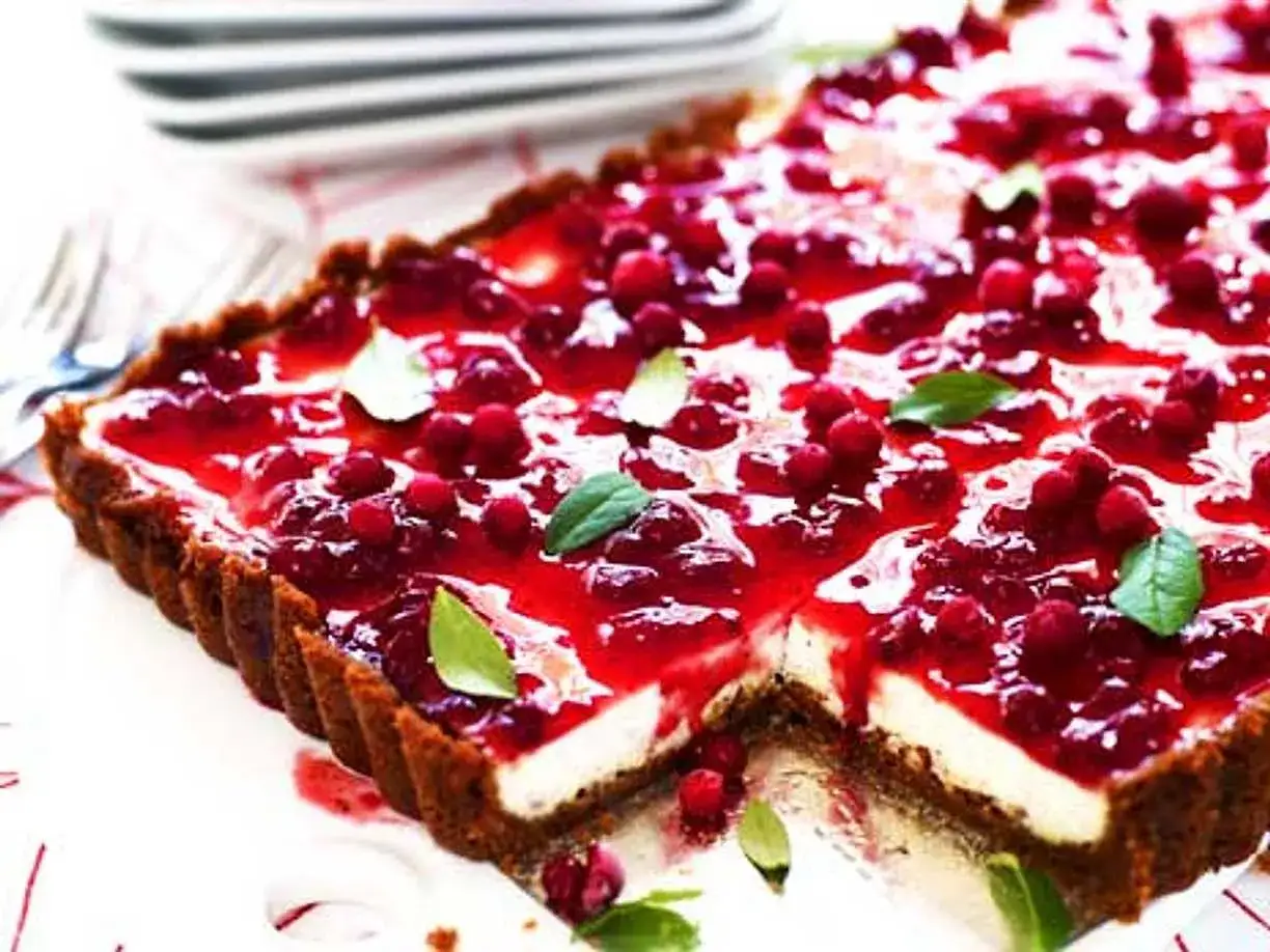 En rektangulär pepparkakscheesecake med en glansig lingon-topping och färska lingon. Garnerad med gröna blad.