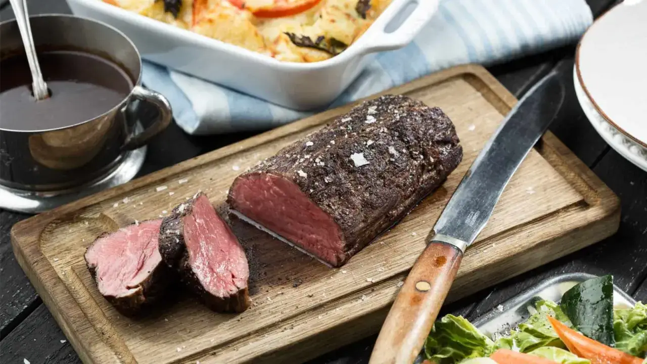 En saftig, helstekt oxfile med flingsalt på en träskärbräda, redo att serveras med en skål sås och en fräsch sallad.