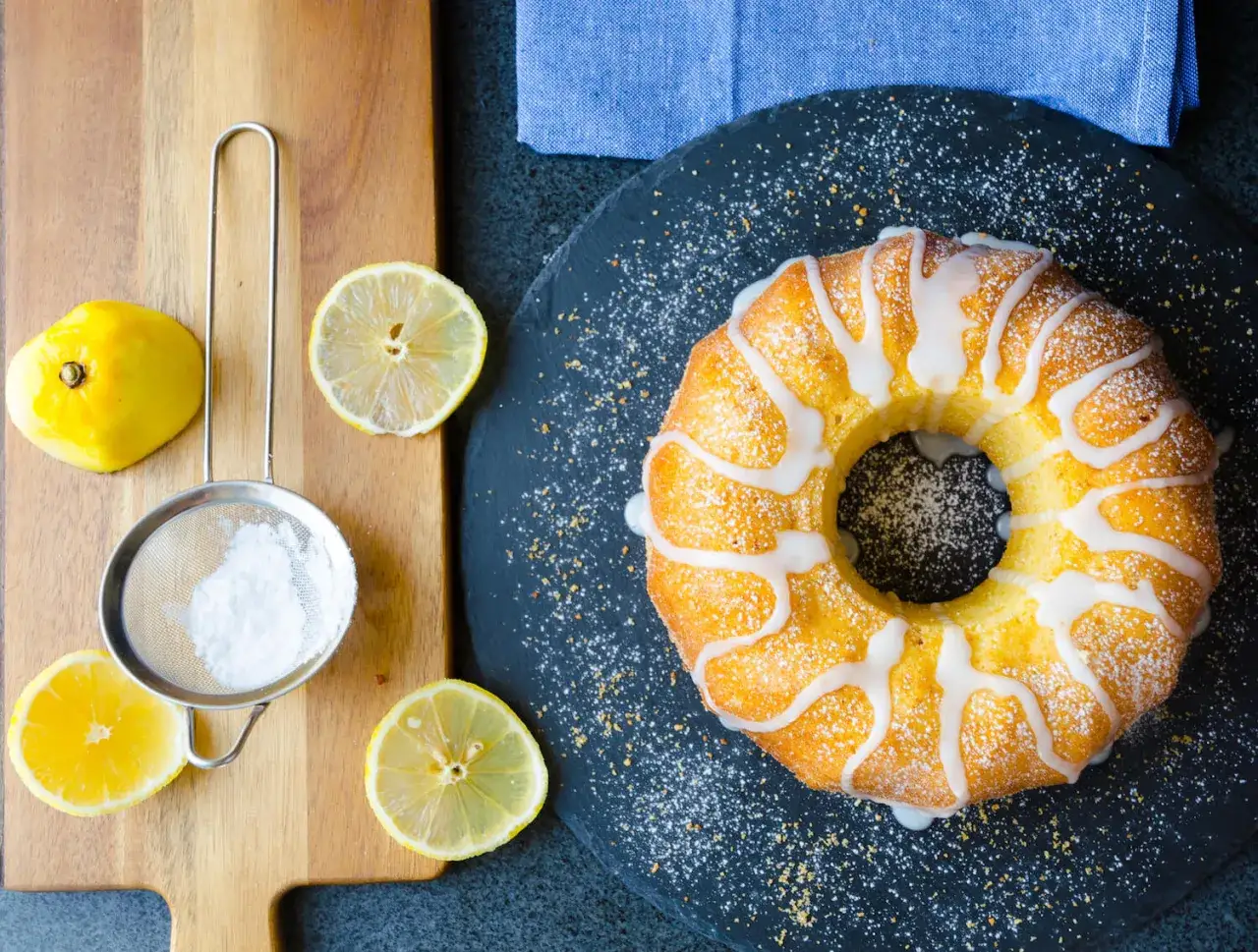 En saftig citron sockerkaka med glasyr och strösocker, omgiven av färska citroner och en sil.
