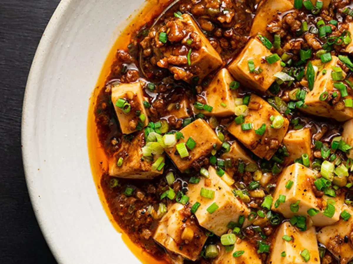 En n&auml;rbild av mapo tofu i en vit sk&aring;l, med mjuka to cubes i en kryddig, k&ouml;ttf&auml;rsbaserad s&aring;s, toppad med f&auml;rsk salladsl&ouml;k.