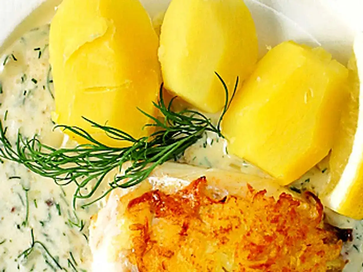 En gyllene fiskbit med krämig sås till fisk, dill och kokta potatisar. En citronskiva ligger vid sidan.
