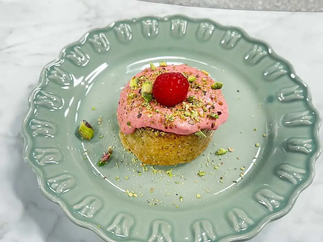 En liten citron kladdkaka med rosa frosting, pistagenötter och ett hallon på en mintgrön tallrik.