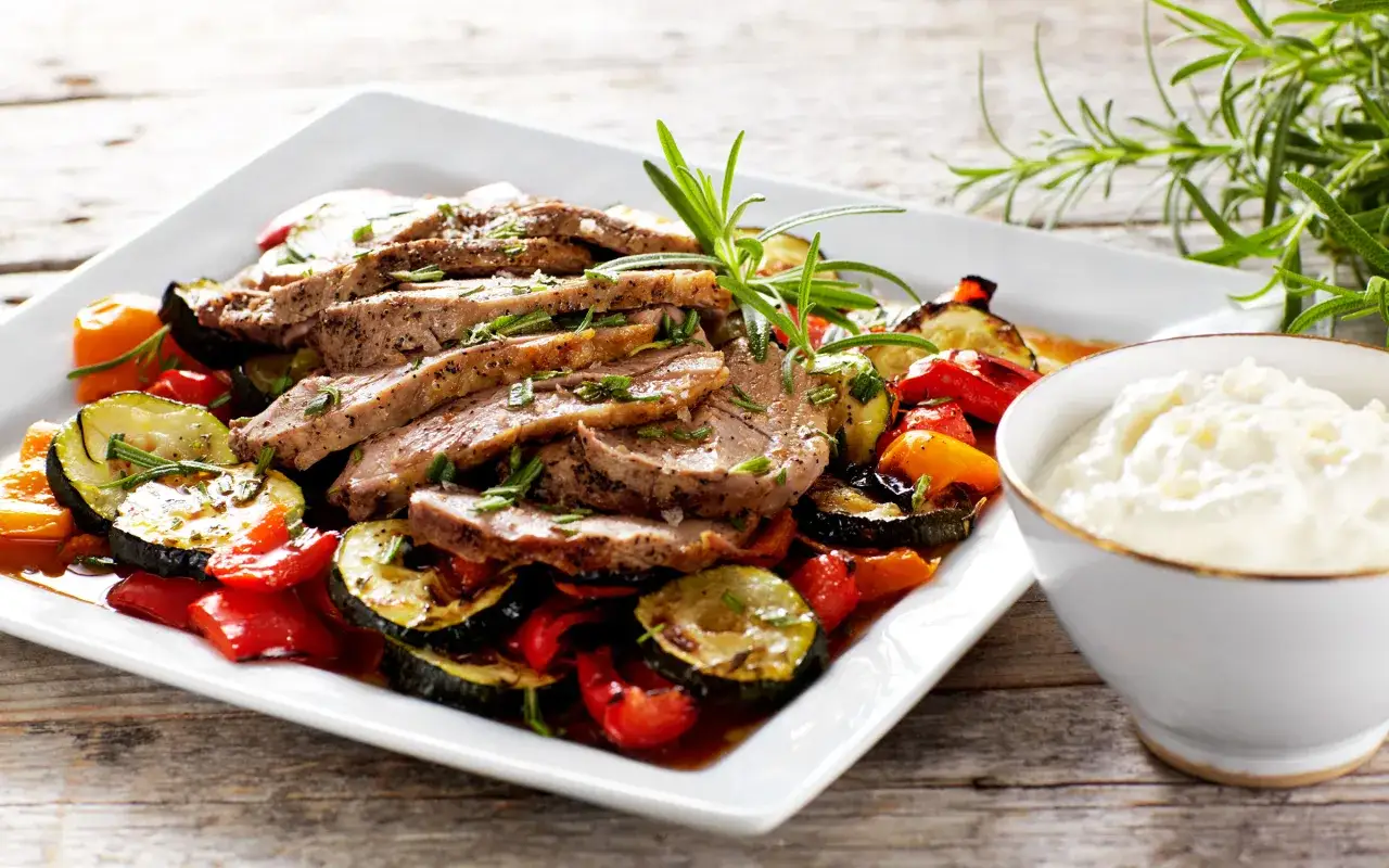 Skivad lammstek i ugn serverad med ugnsrostade grönsaker som zucchini, paprika och tomater. En klick vit sås i en skål.