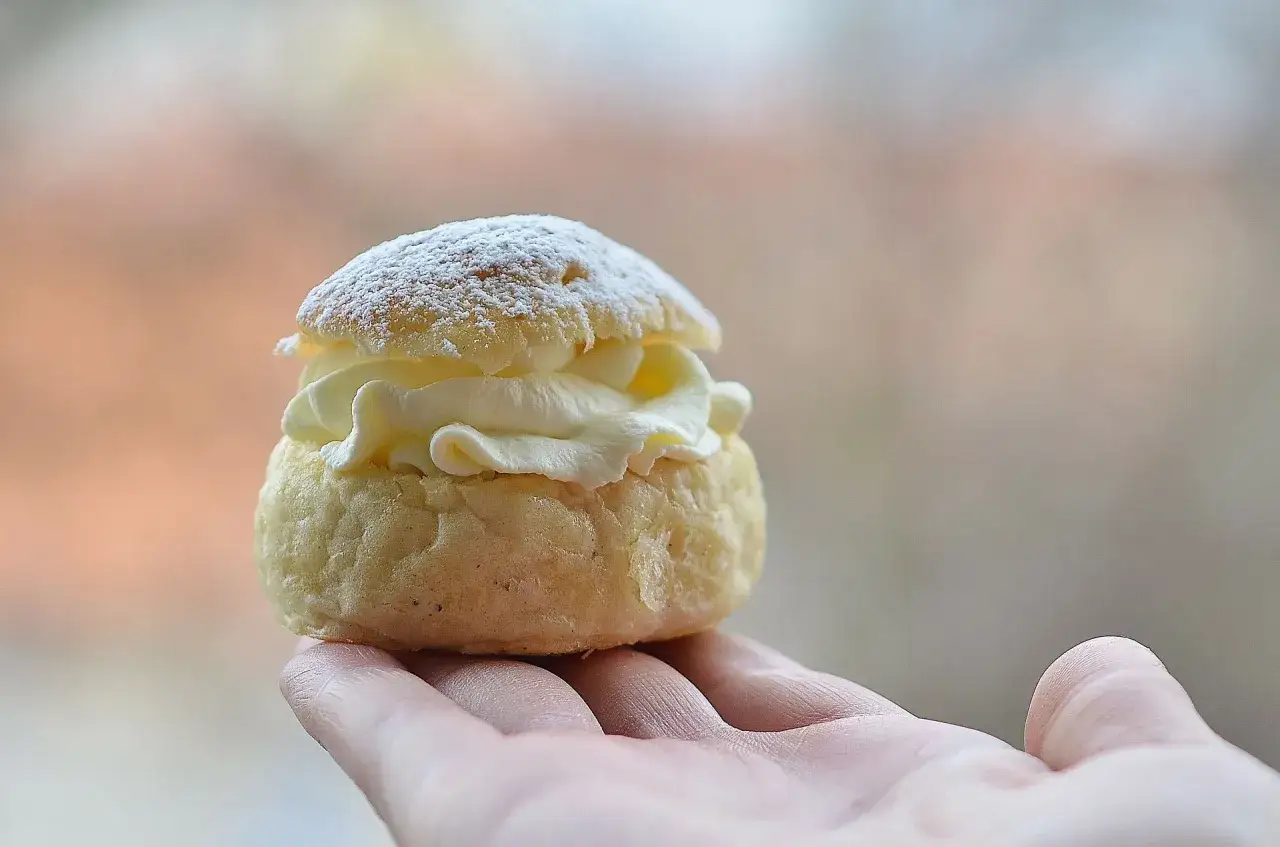 En hand håller en nybakad semla, pudrad med florsocker. Perfekt för att baka semlor till fikat.