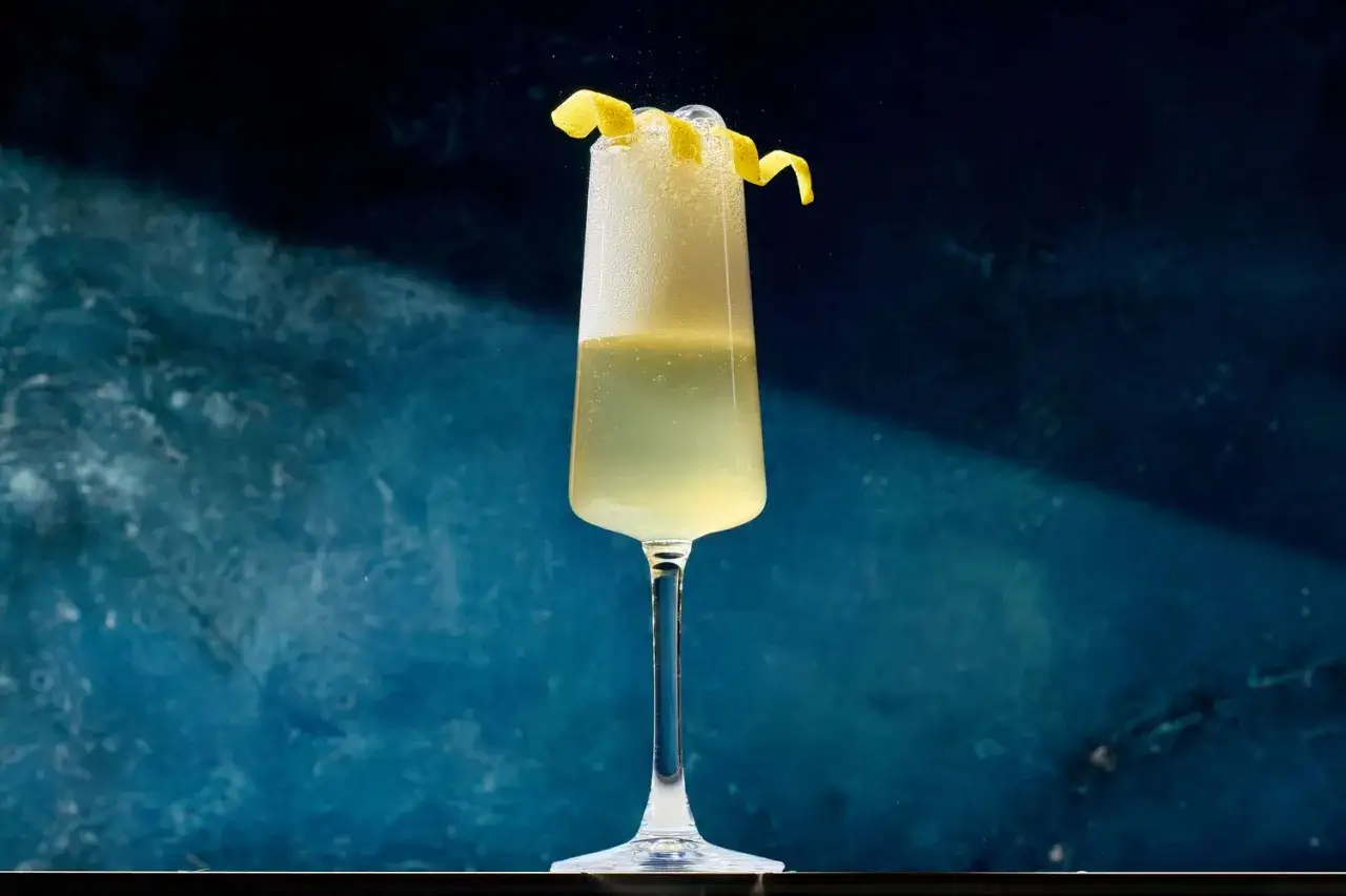 Ett elegant glas med en French 75, toppad med citronskal och bubblor, mot en m&ouml;rkbl&aring; bakgrund.