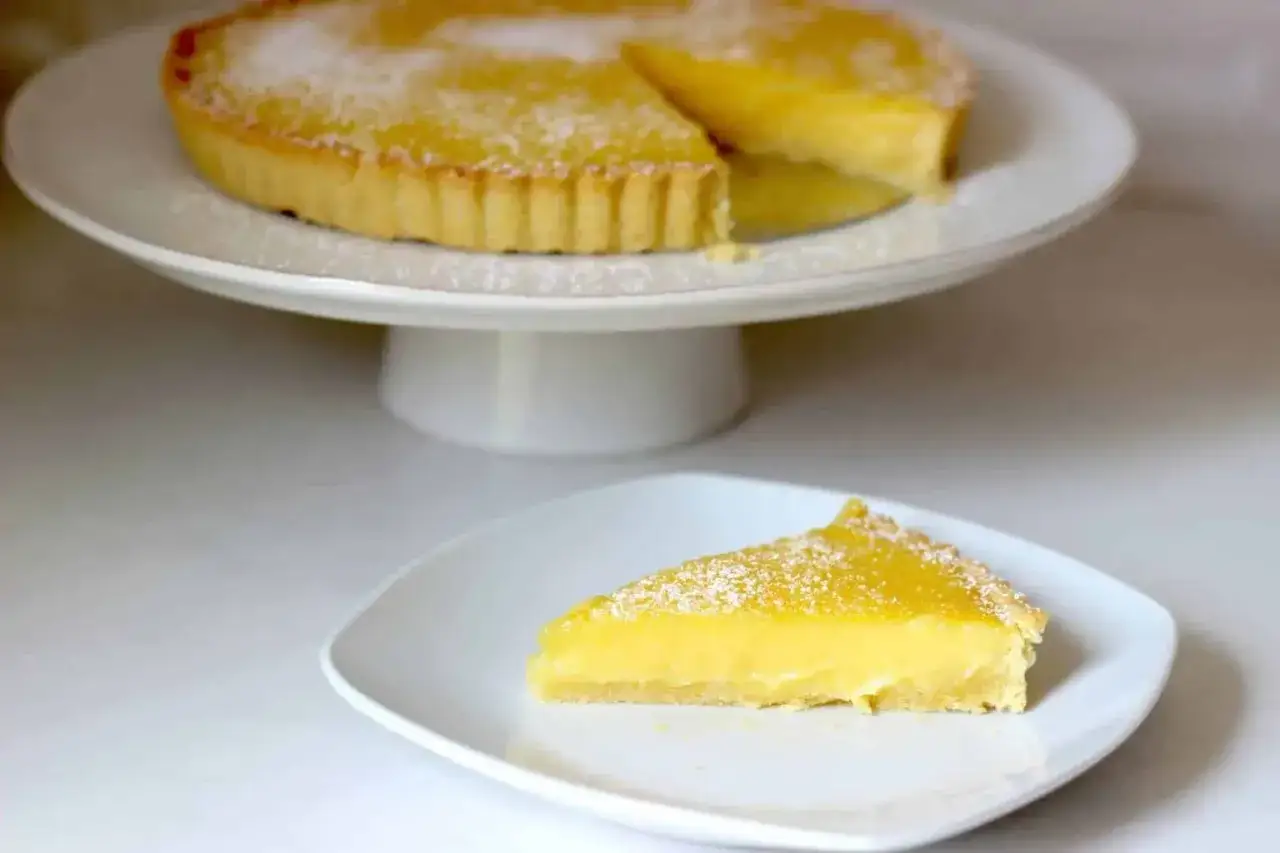 En härlig citron tarte med en krämig fyllning och pudrad florsocker. En bit av den syrliga godsaken ligger på en vit tallrik.