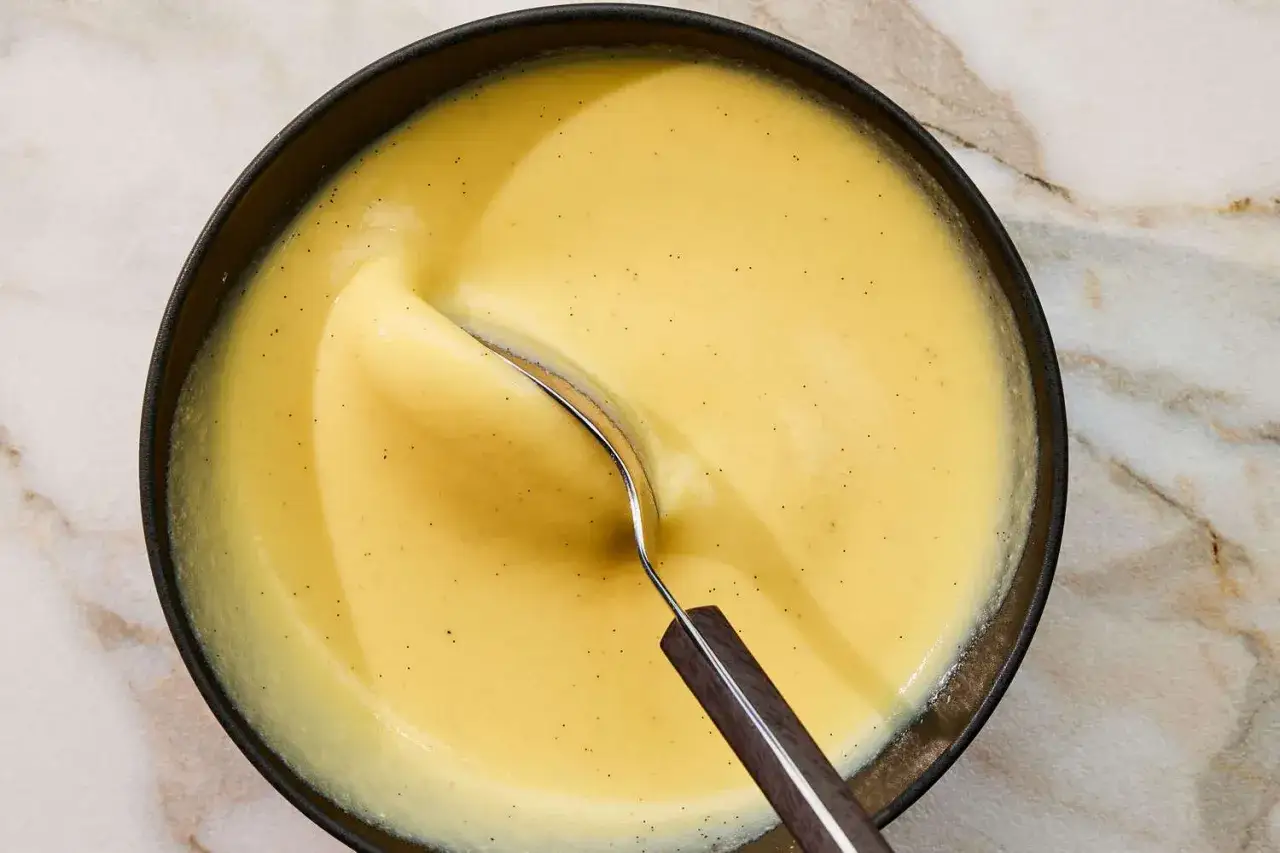 En skål med krämig vaniljsås, creme anglaise, med små prickar av vanilj. En sked vilar i den lena krämen.