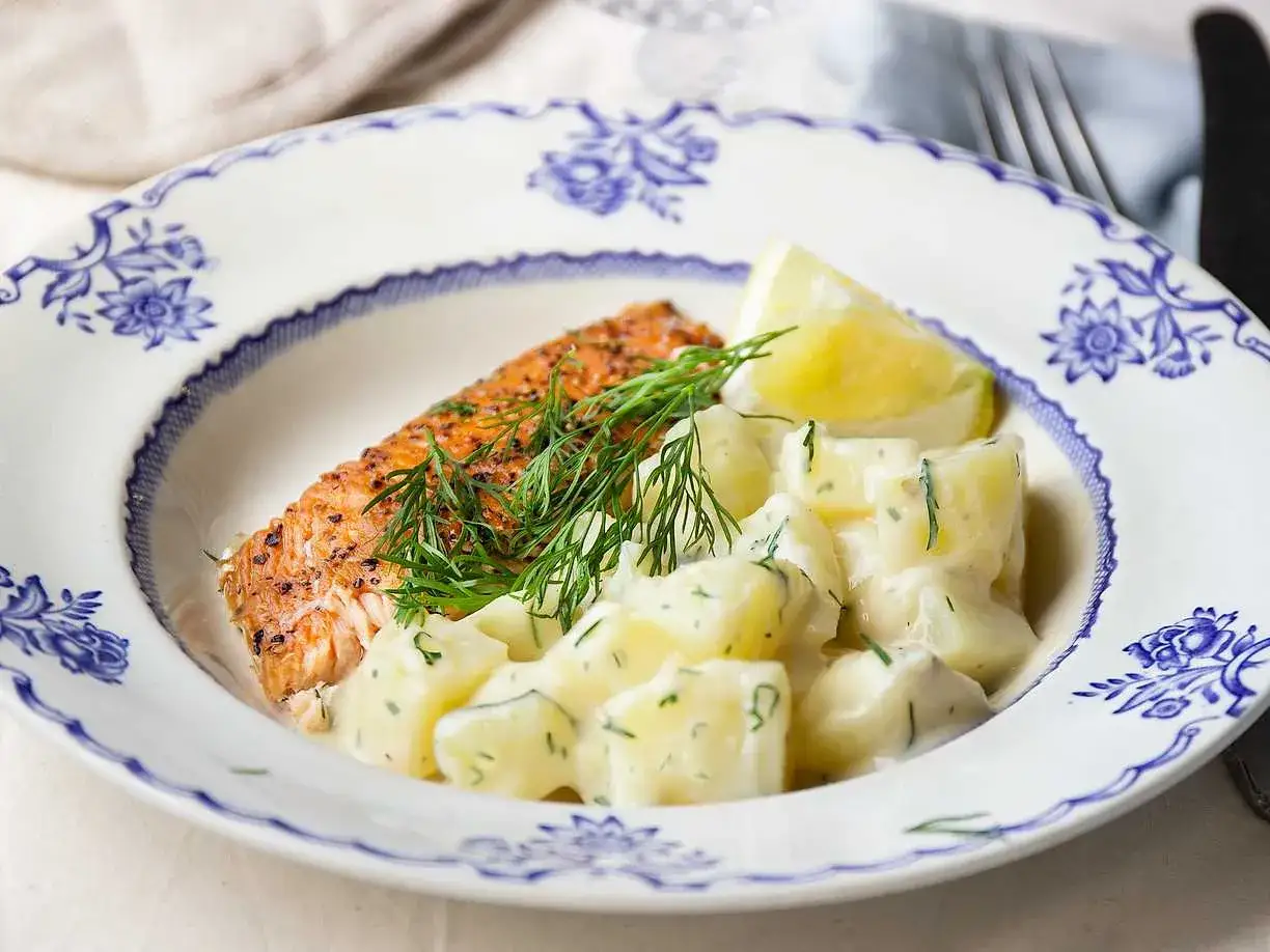 En saftig laxfilé serveras med krämig dillstuvad potatis och en citronklyfta på en vacker blåvit tallrik.