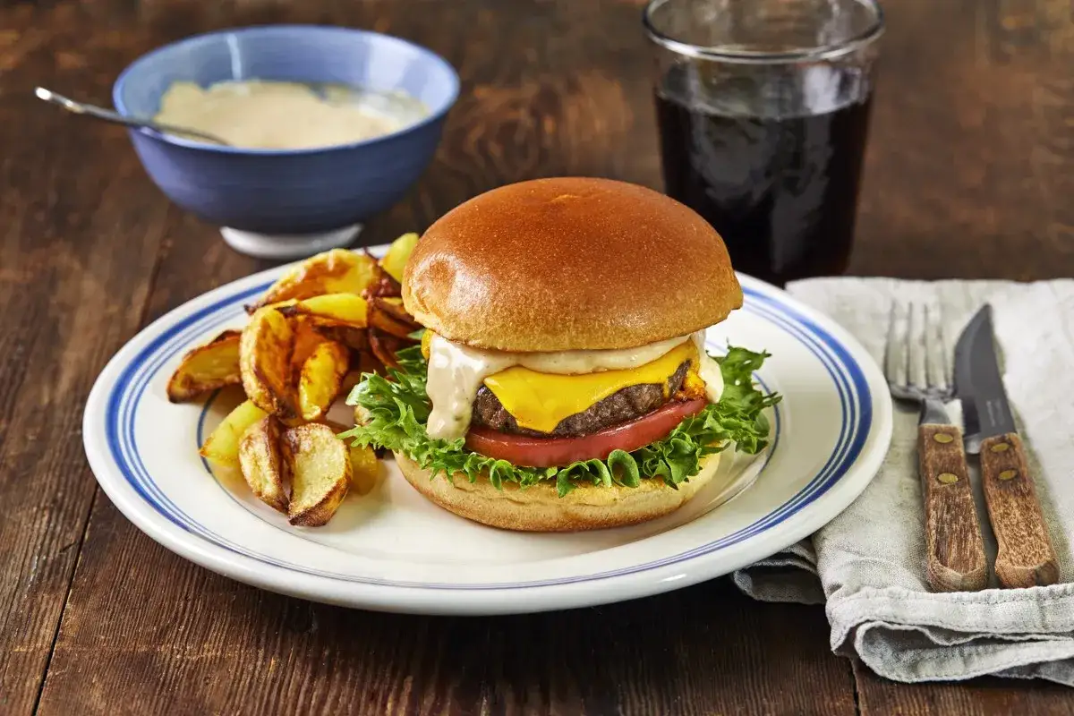 En saftig hamburgare med ost, sallad och tomat, serverad med klyftpotatis och en sk&aring;l med hemlagad hamburgerdressing.