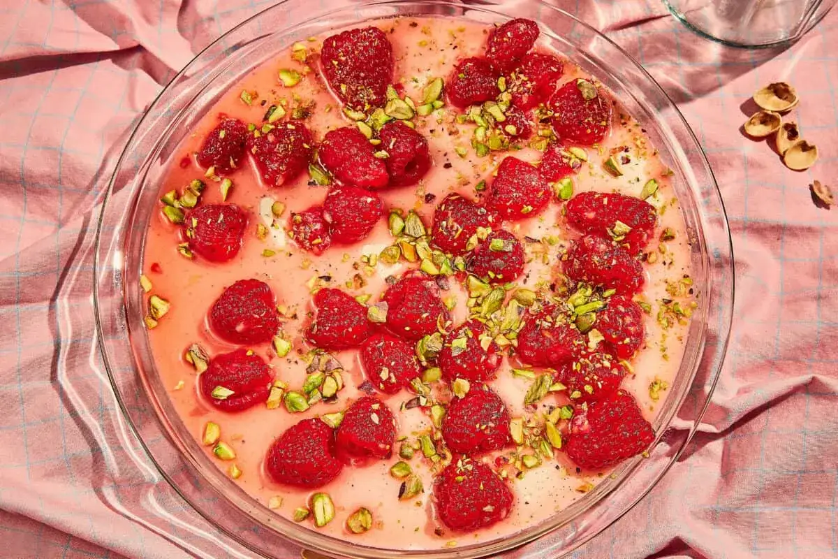 En kr&auml;mig panna cotta med hallon och pistagen&ouml;tter i en glasform, serverad p&aring; en rosa duk.