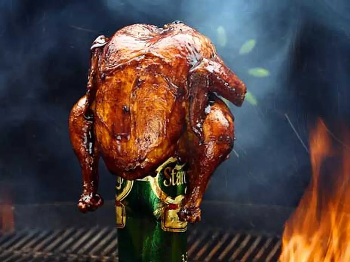 En saftig ölkyckling, perfekt grillad med gyllene skinn, står stolt på en ölburk. Flammor och rök skapar en dramatisk bakgrund.