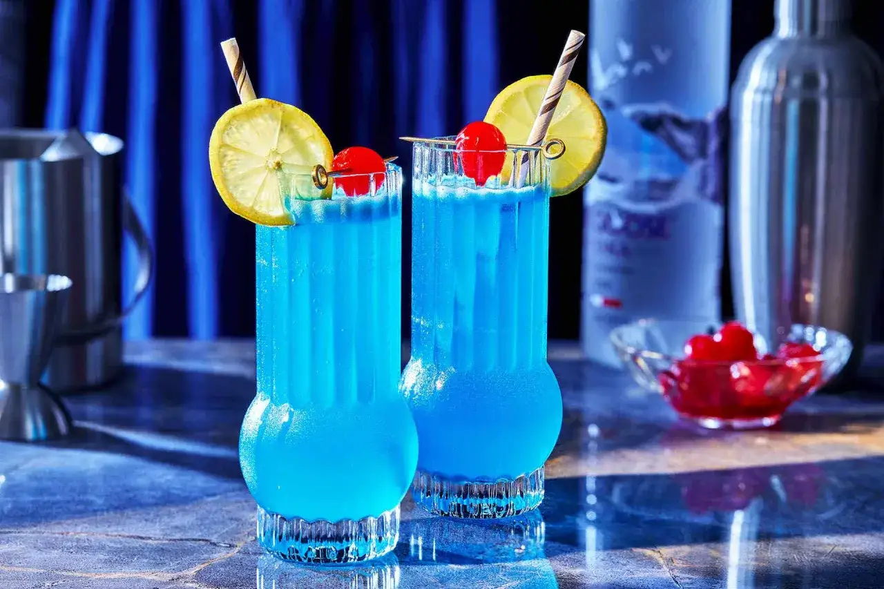 Tv&aring; bl&aring; drinkar, en bl&aring; lagun, med citron och k&ouml;rsb&auml;r. En flaska vodka och en shaker i bakgrunden.