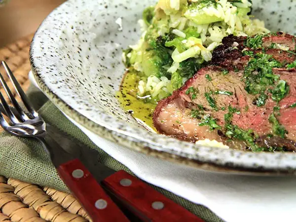 Close-up of grilled picanha steak with rice and greens, served on a speckled plate with a fork and knife.