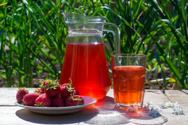 En karaff och ett glas med röd saft står på ett träbord. En tallrik med färska jordgubbar bredvid. Perfekt för ett enkelt jordgubbssaft recept.