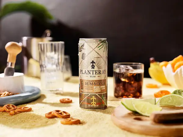 En burk Planteray Rum & Cola står på ett gult underlag, bredvid ett glas med is och mörk dryck. Små pretzels och limeklyftor ligger utspridda.