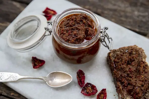 En burk med soltorkad tomat-tapenade, perfekt för ett enkelt tapenade recept. Serveras med en skiva mörkt bröd och en silver sked.