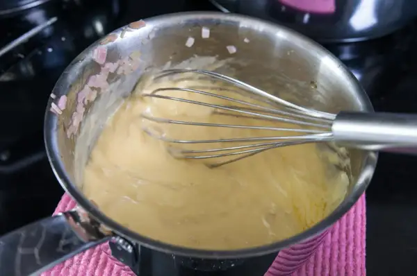 En visp rör i en gul sås i en kastrull. Små bitar av schalottenlök syns på kastrullkanten. Ett perfekt bearnaisesås recept börjar här.