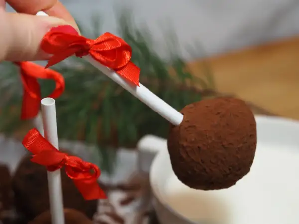 En hand håller en chokladboll på pinne med röd rosett, redo att doppas i en mugg med varm choklad.