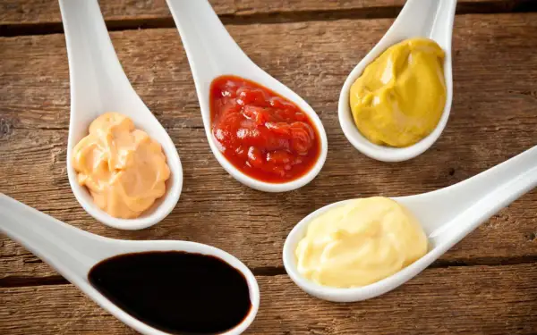 Fem skedar med olika såser: ketchup, majonnäs, senap, en orange sås och mörk bbq sås på träbakgrund.
