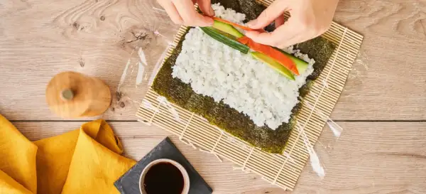 Händer lägger lax och gurka på ris för ett sushi recept.